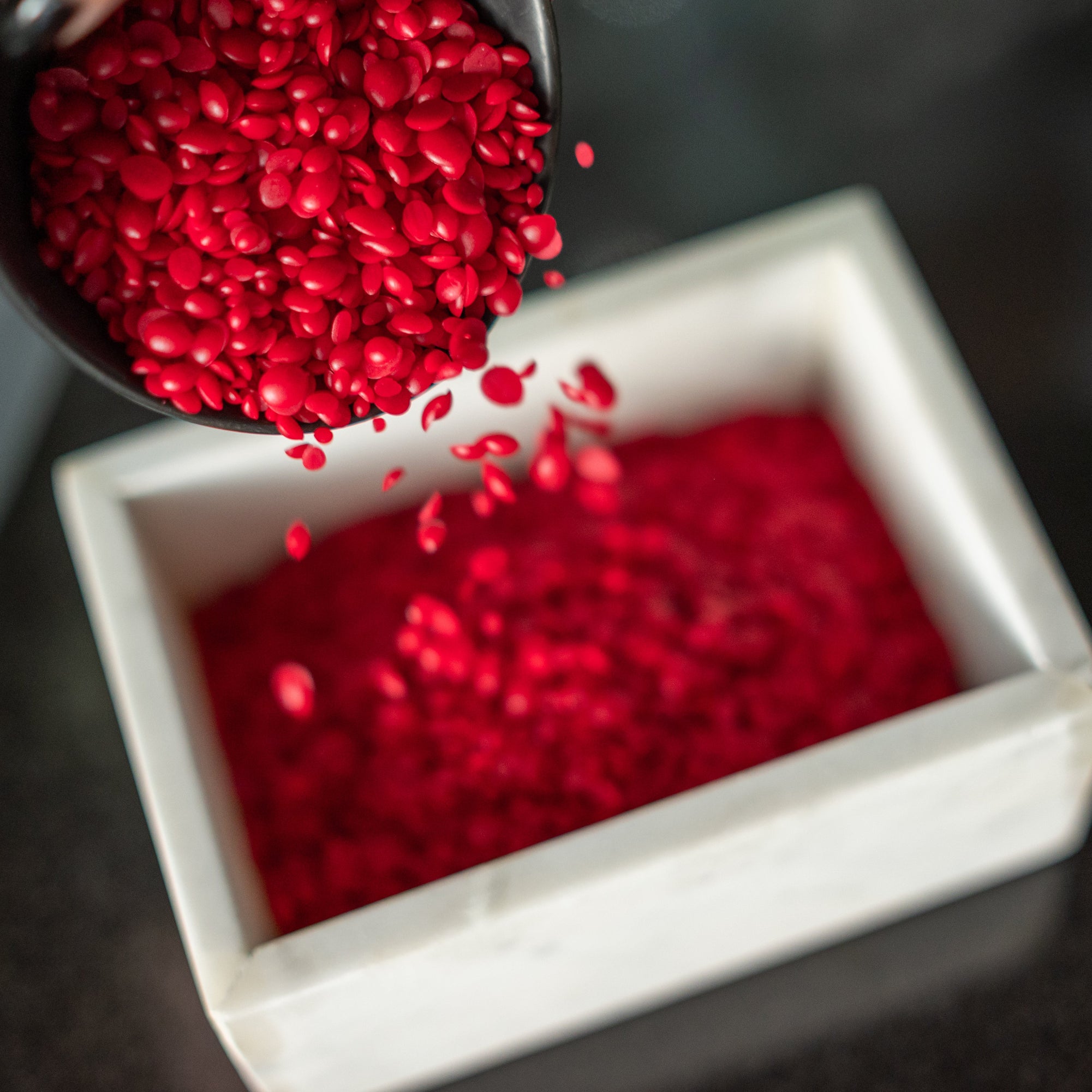 Red wax being poured from a metal scoop into a white container on a dark surface.